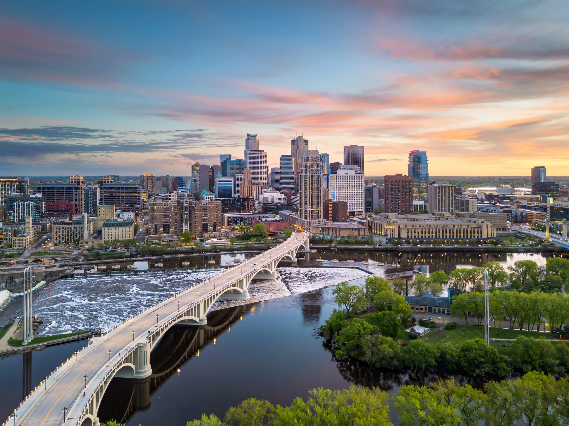 The Minneapolis skyline as the sun sets, showing a bridge in the foreground and buildings beyond.