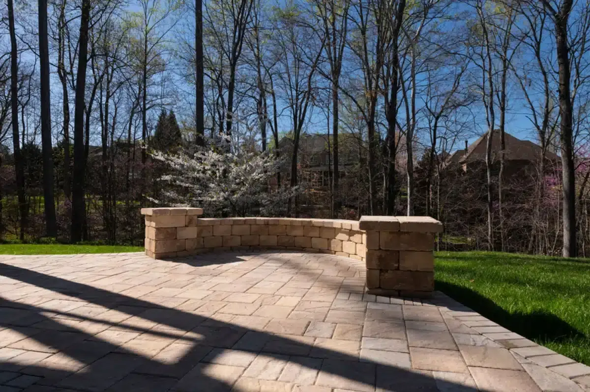 A beige colored paver patio with matching seating wall and woods beyond
