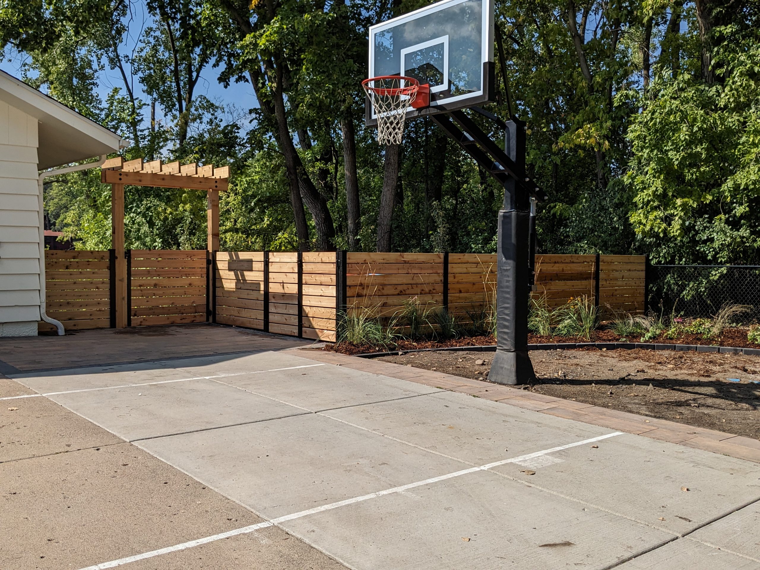 A newly built backyard basketball court with a wooden fence and pergola, surrounded by fresh landscaping.