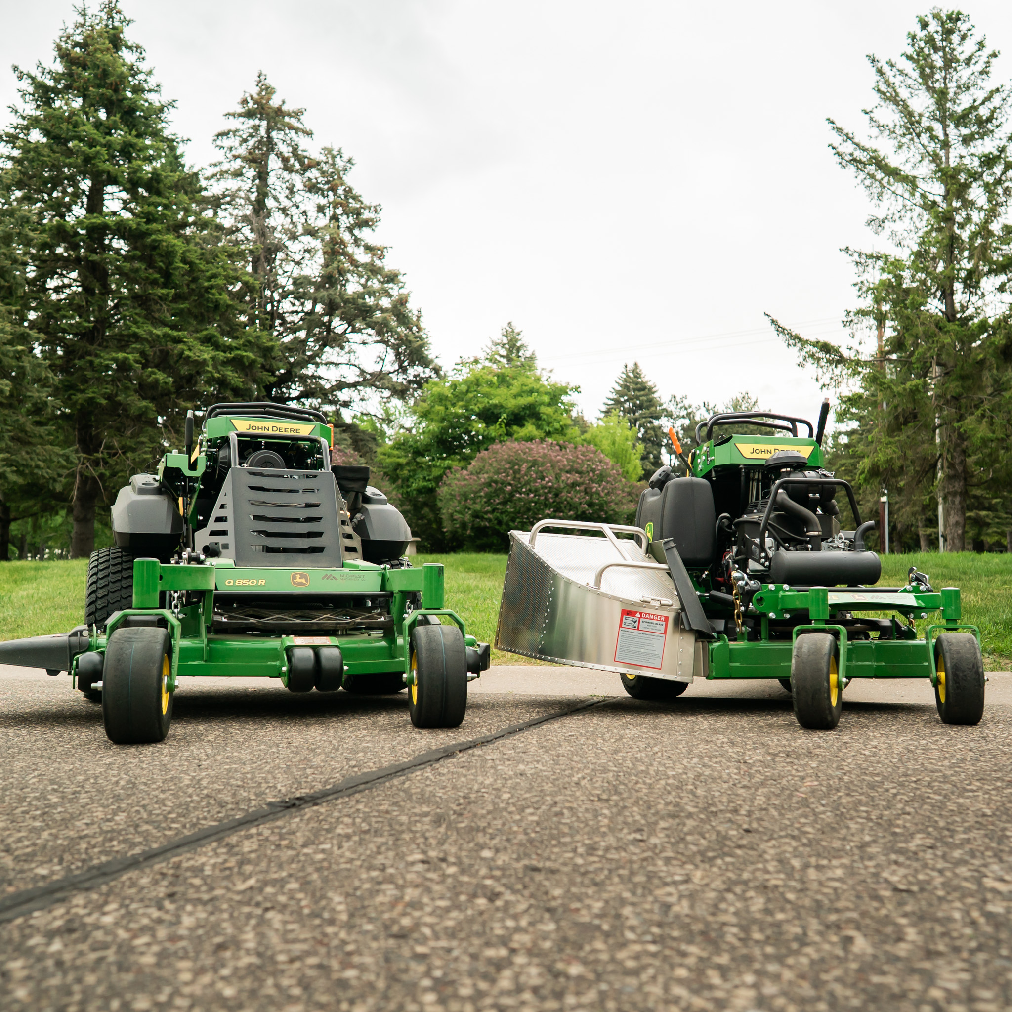 Two John Deere lawn mowers parked on gravel with green grass in the background.