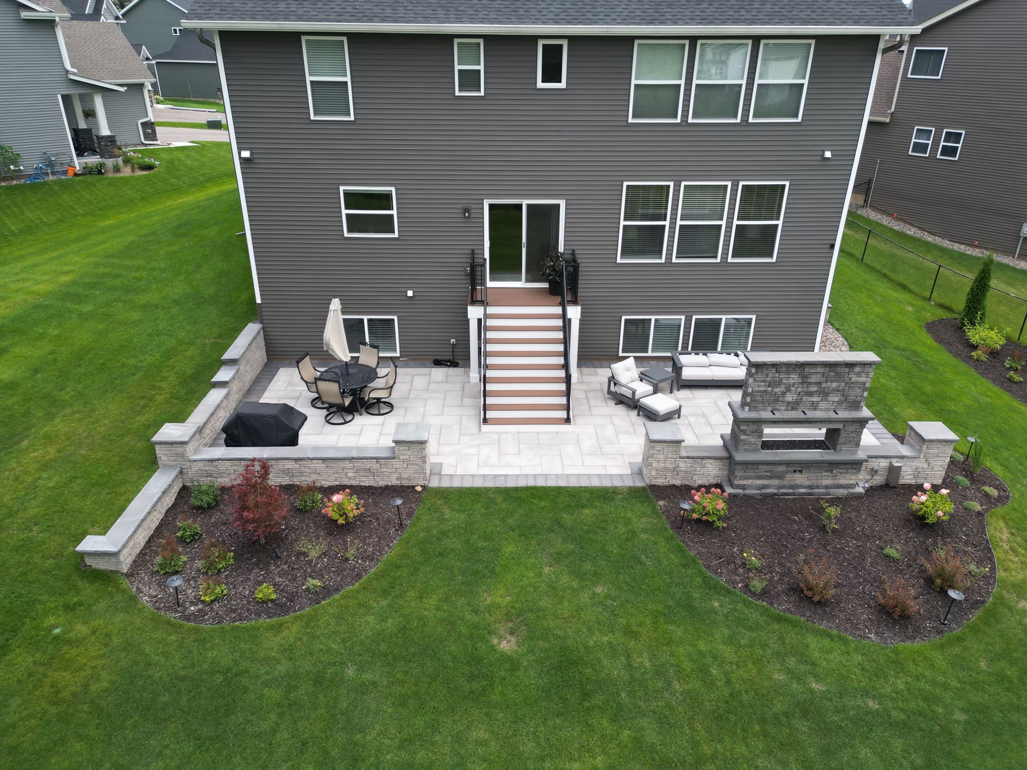 A landscaped backyard with a stone patio, built-in fire pit, seating wall, and flower beds beside a green lawn.