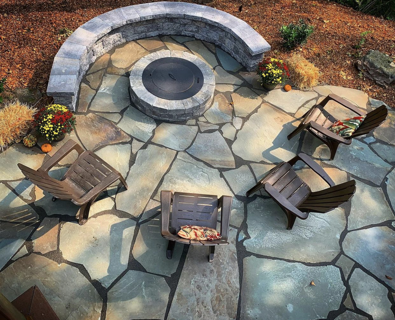 A circular stone fire pit surrounded by a stone seating wall and wooden chairs on a stone patio.