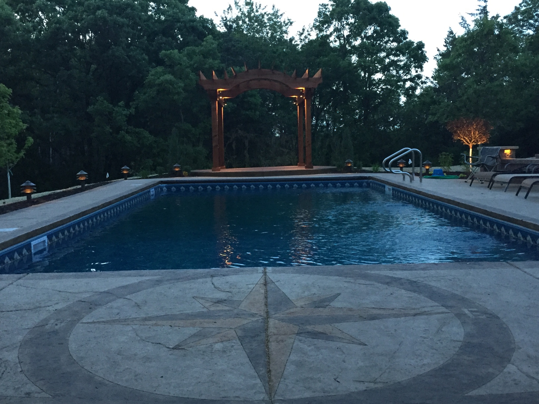A concrete swimming pool with a stamped concrete patio and a wooden pergola, lit for an inviting evening atmosphere.